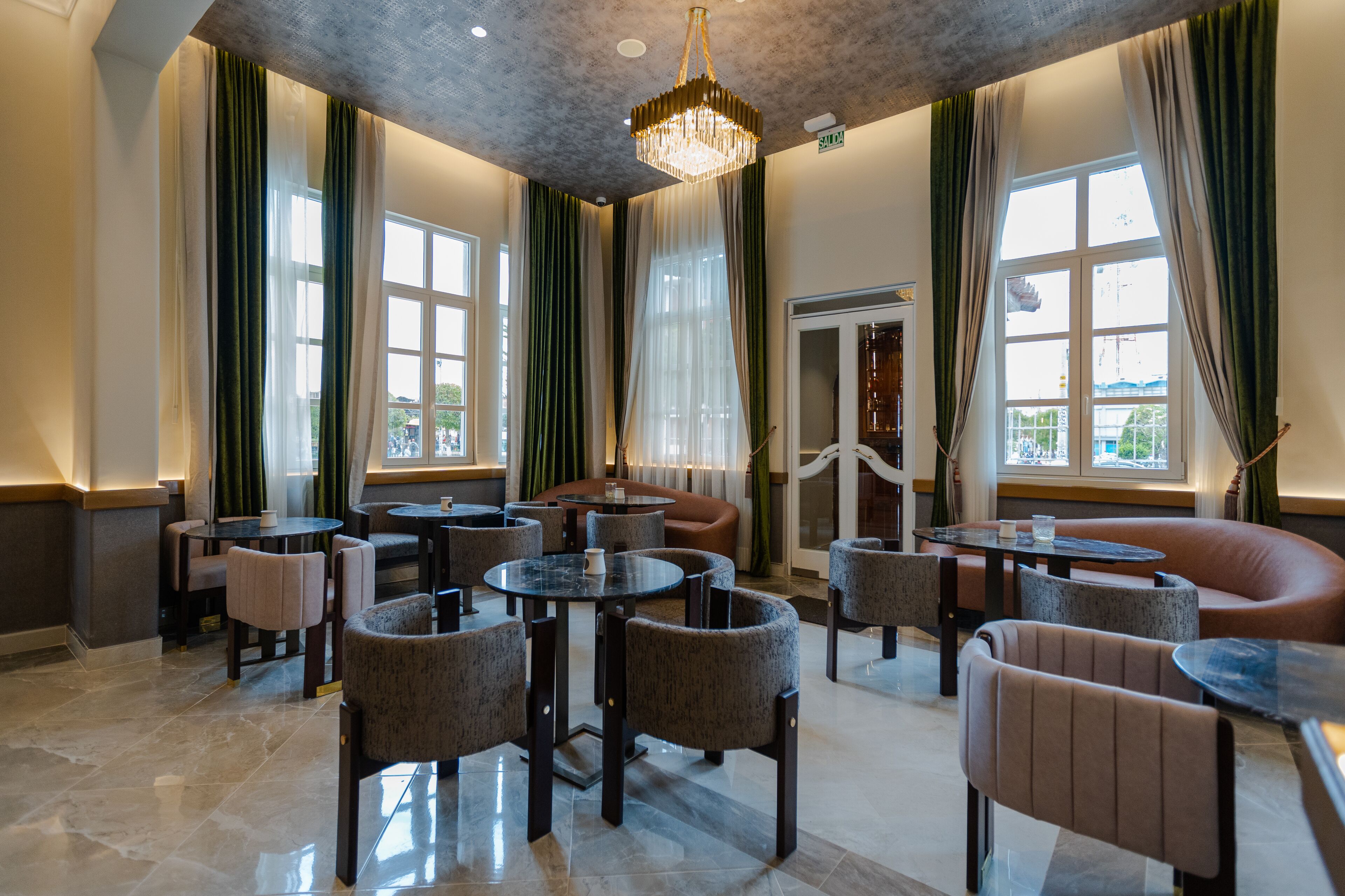 Eine Hotelhalle mit runden Tischen und Stühlen, großen Fenstern mit Vorhängen und einer Kristallleuchter.
