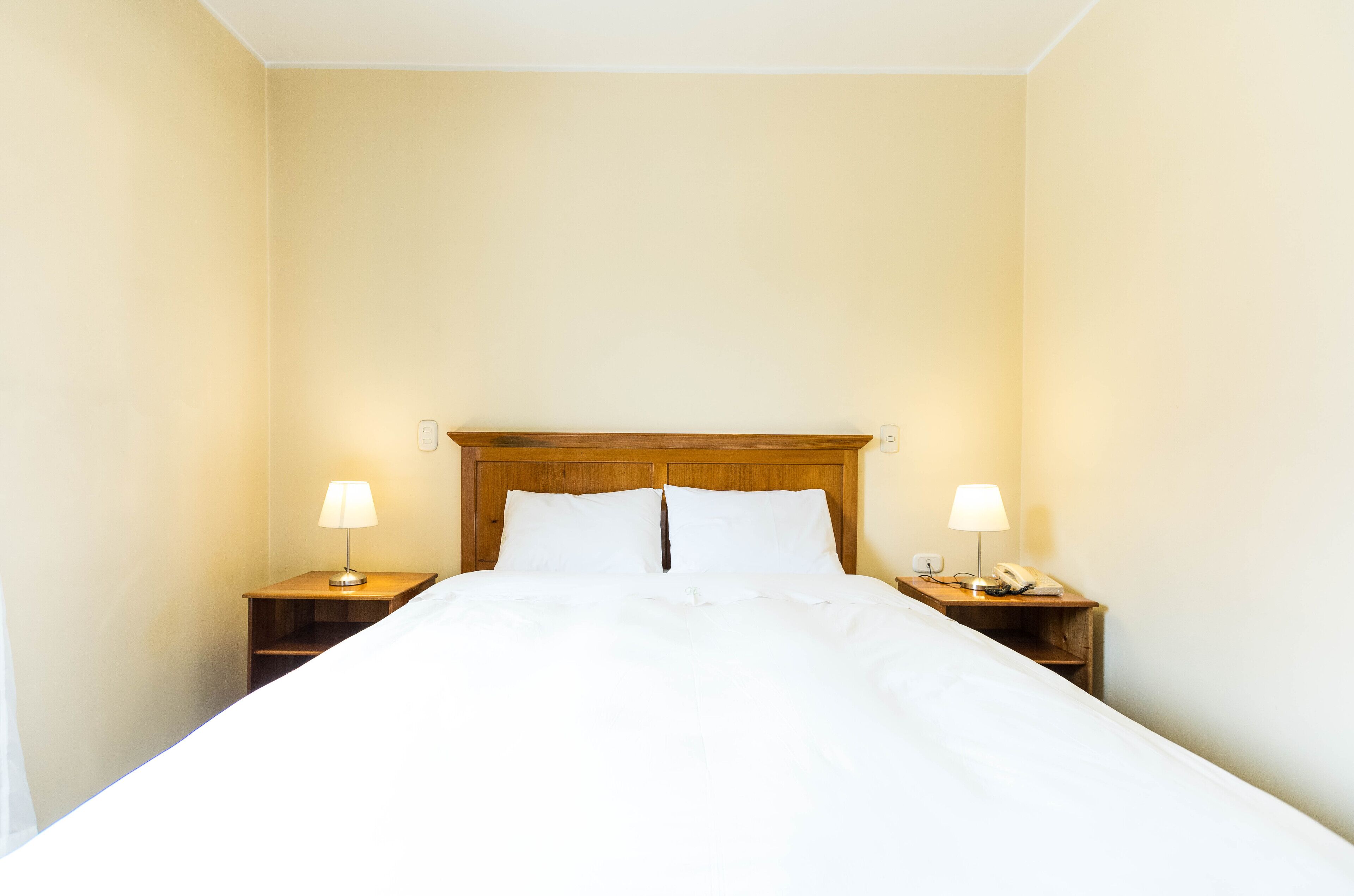 Una cama bien hecha con sábanas blancas, dos mesitas de noche con lámparas y un cabecero de madera.