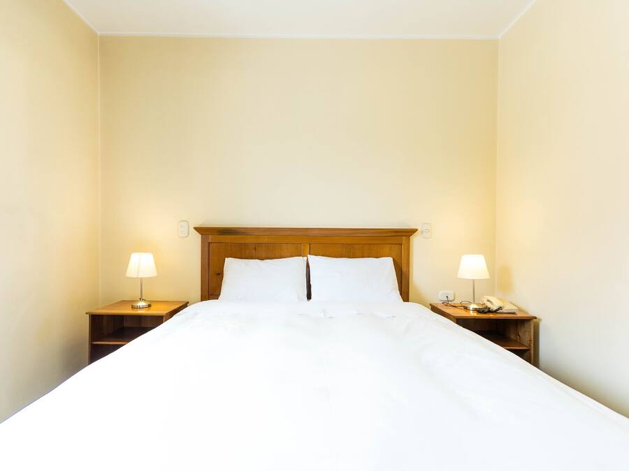 Una cama bien hecha con sábanas blancas, dos mesitas de noche con lámparas y un cabecero de madera.