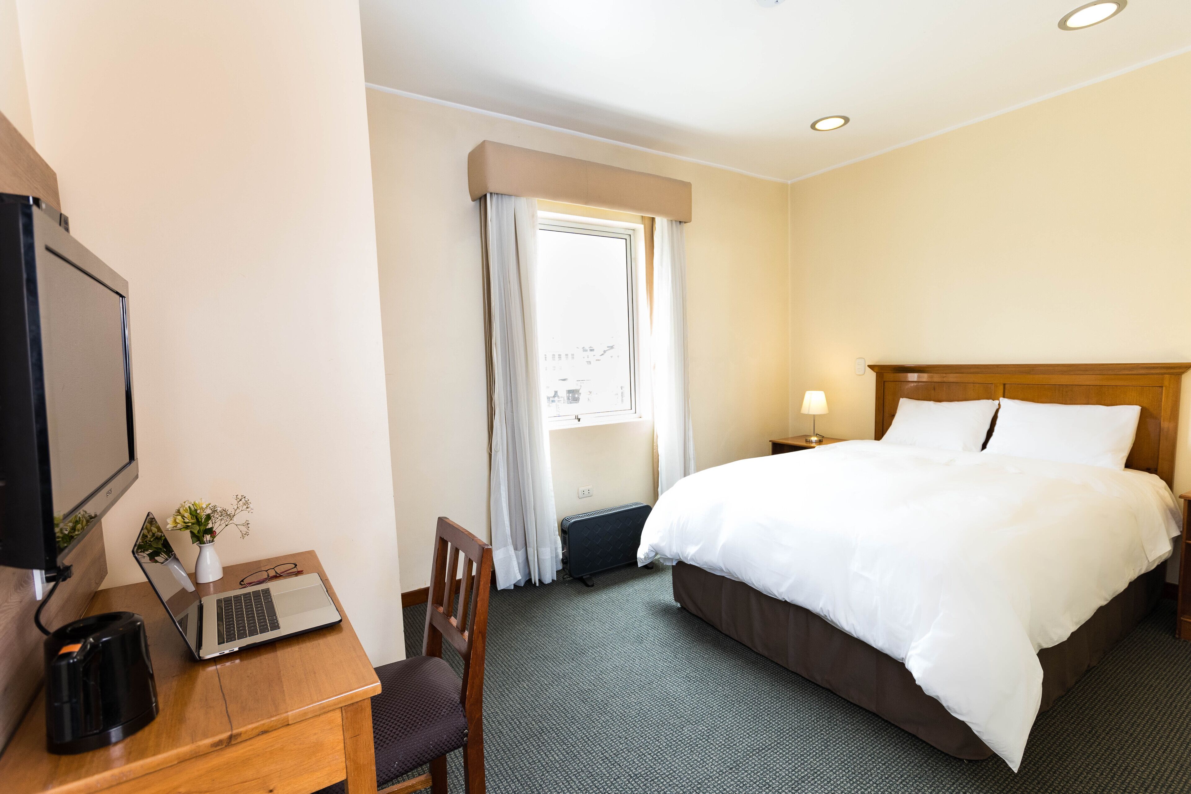 Une chambre d’hôtel avec un lit, un bureau avec un ordinateur portable, une chaise, une télévision et une fenêtre avec des rideaux.