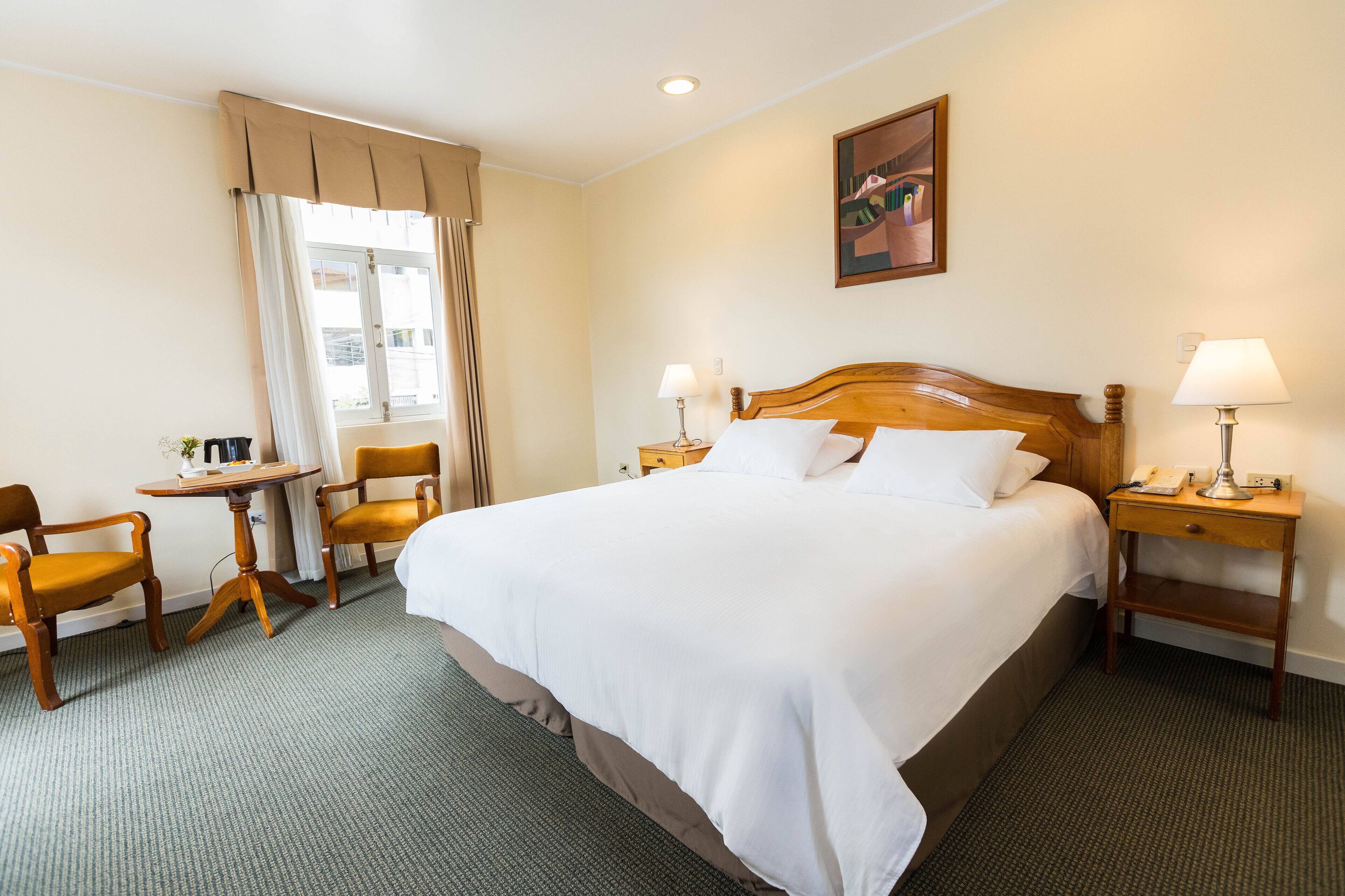A hotel room with a large bed, two bedside tables with lamps, a small table with a chair, and a painting on the wall.