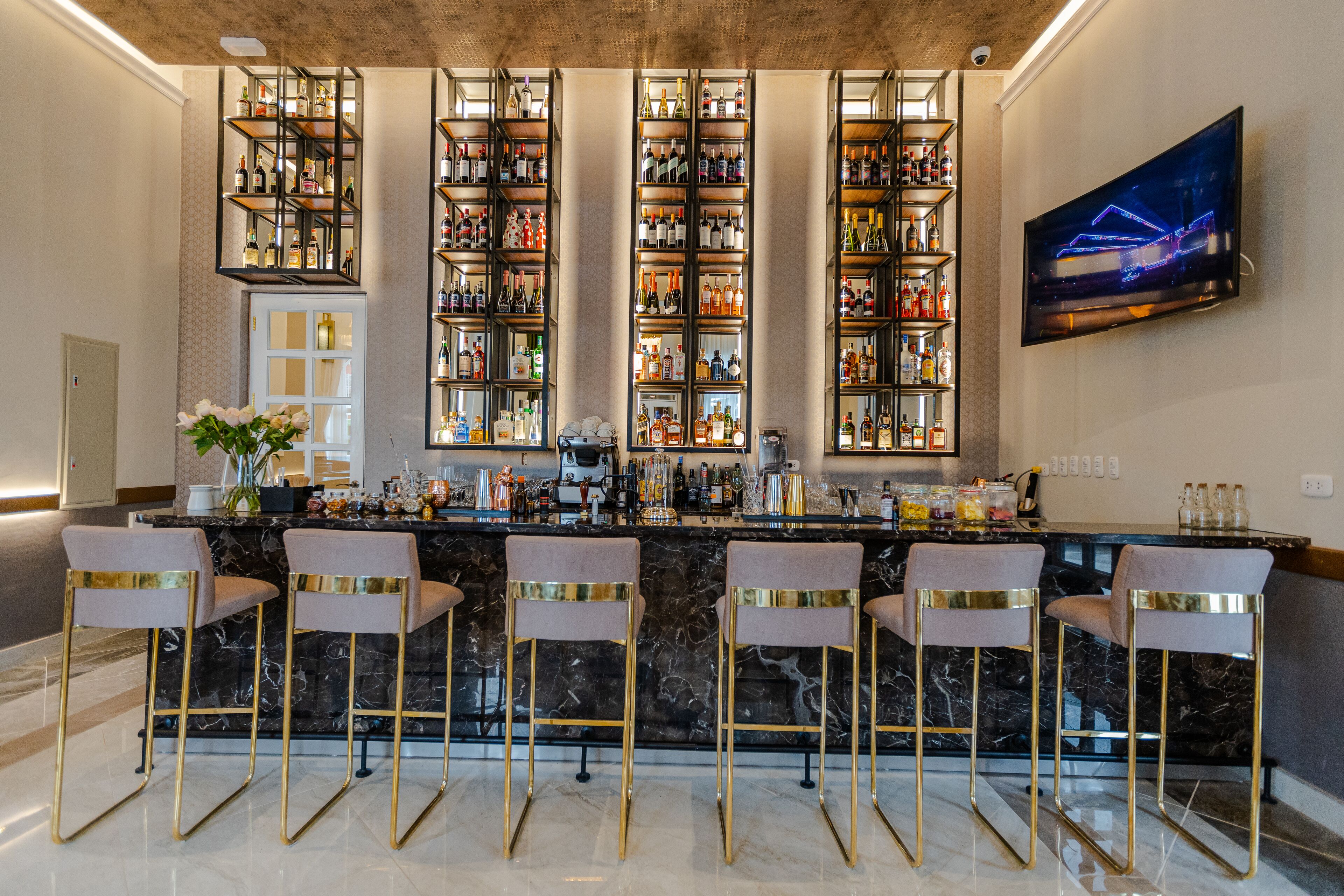 Una zona de bar con mostradores de mármol, varias estanterías llenas de botellas y cuatro taburetes.