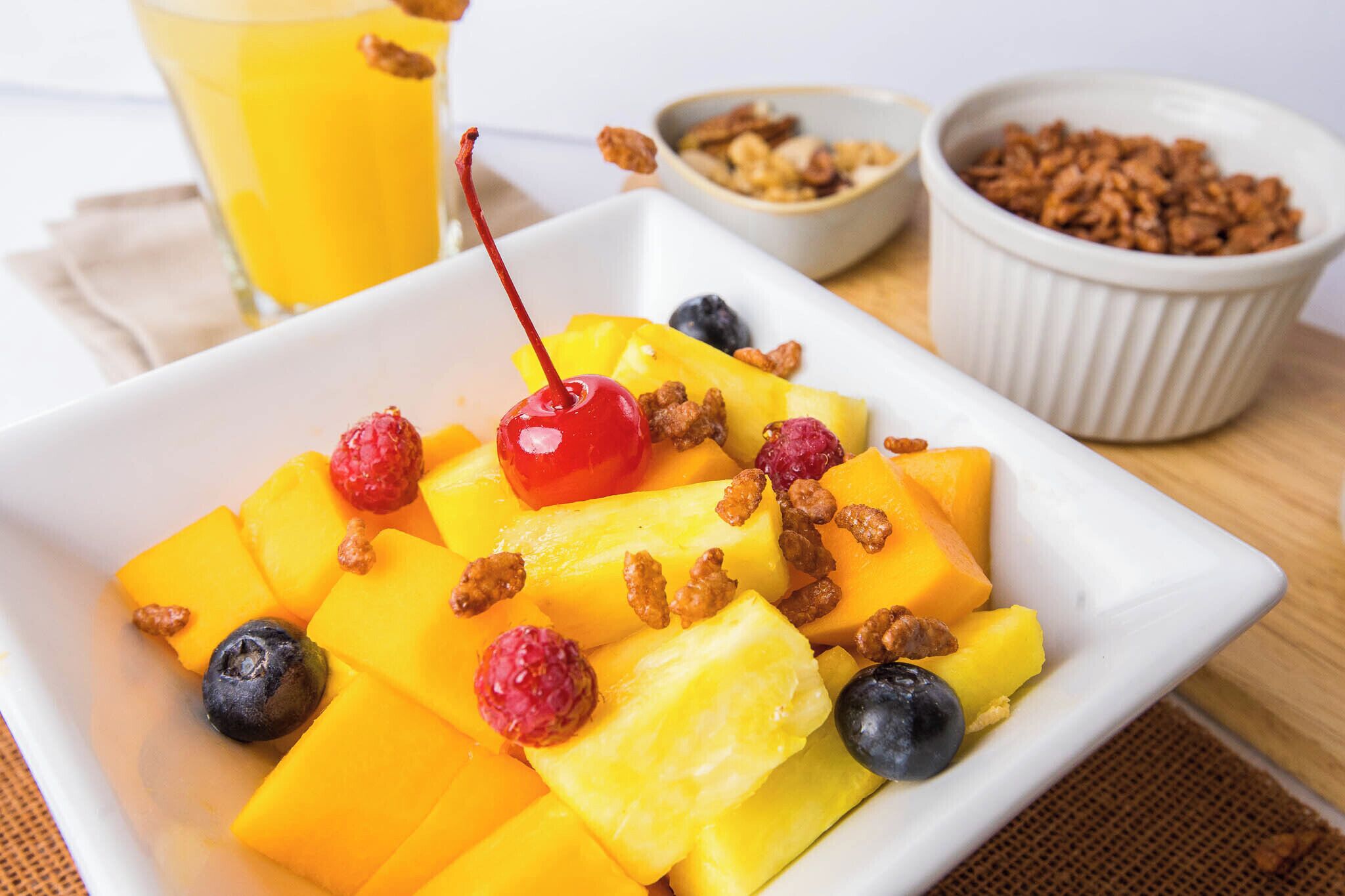 A white square plate with yellow mango chunks, blueberries, raspberries, and a cherry, accompanied by bowls of granola and a glass of orange juice.