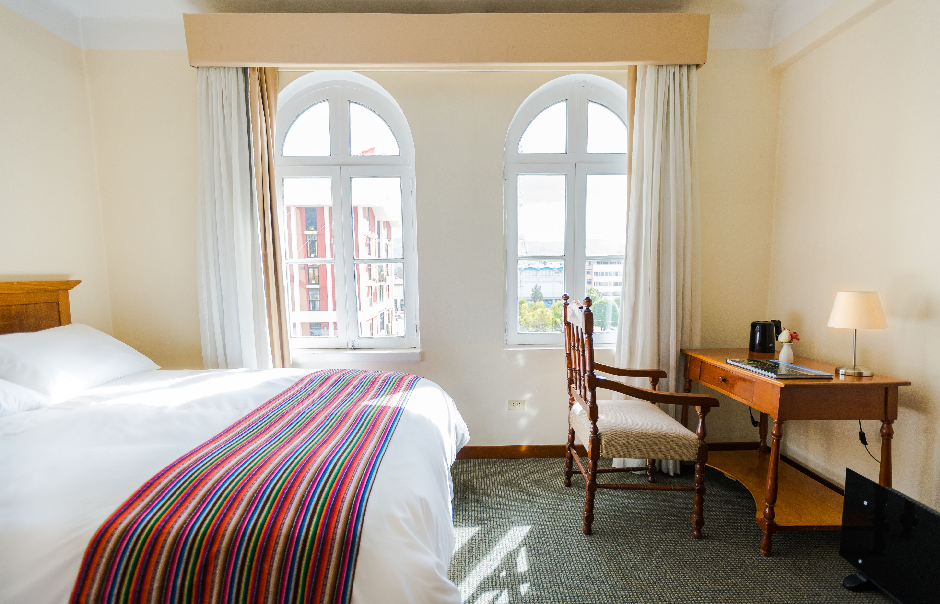 A hotel room with two beds, a desk, a chair, a TV, and large windows with curtains.