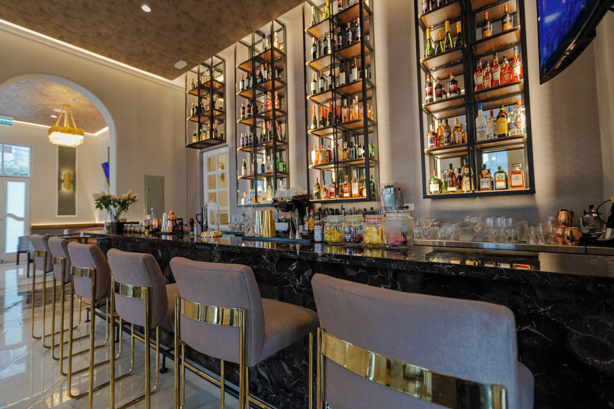 A bar area with multiple shelves of bottles, bar stools with gold accents, and a marble countertop.