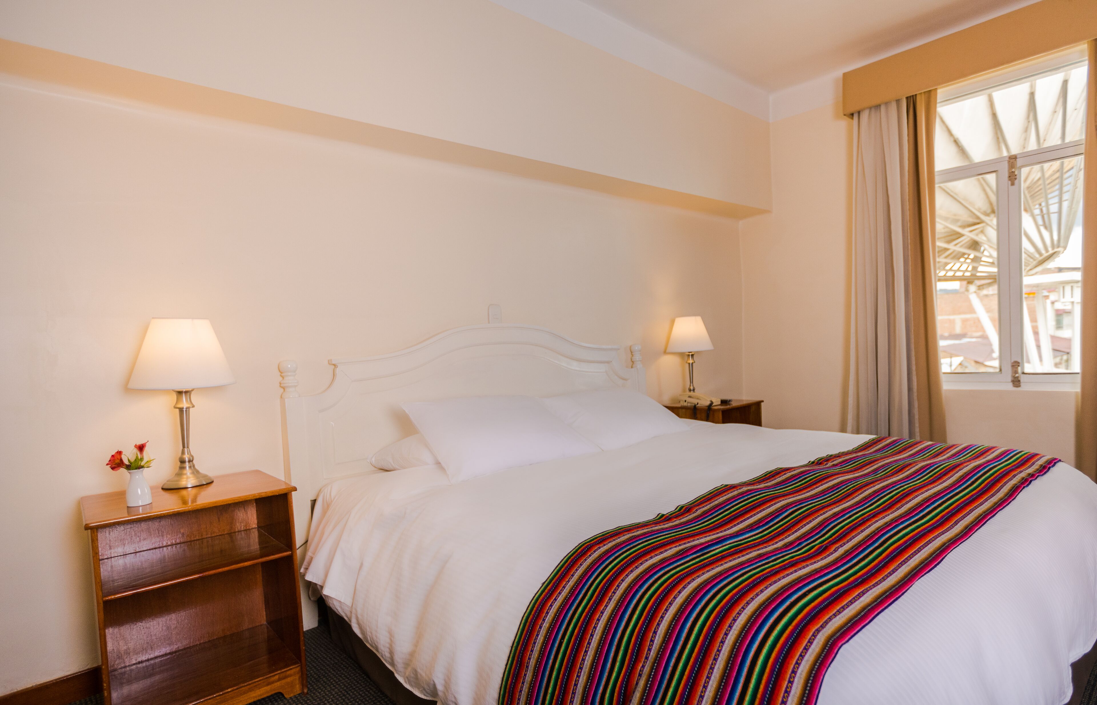 A hotel room with a large bed, two bedside tables with lamps, a wooden nightstand, and a window with curtains.