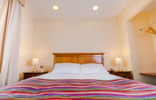 Habitación de hotel con cabecera de madera, dos mesitas de noche con lámparas, una colcha de rayas de colores y una cortina blanca.