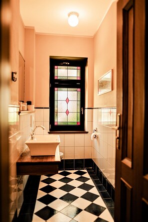 Un baño con piso a cuadros blanco y negro, un lavamanos blanco y una ventana con diseño de vitral.