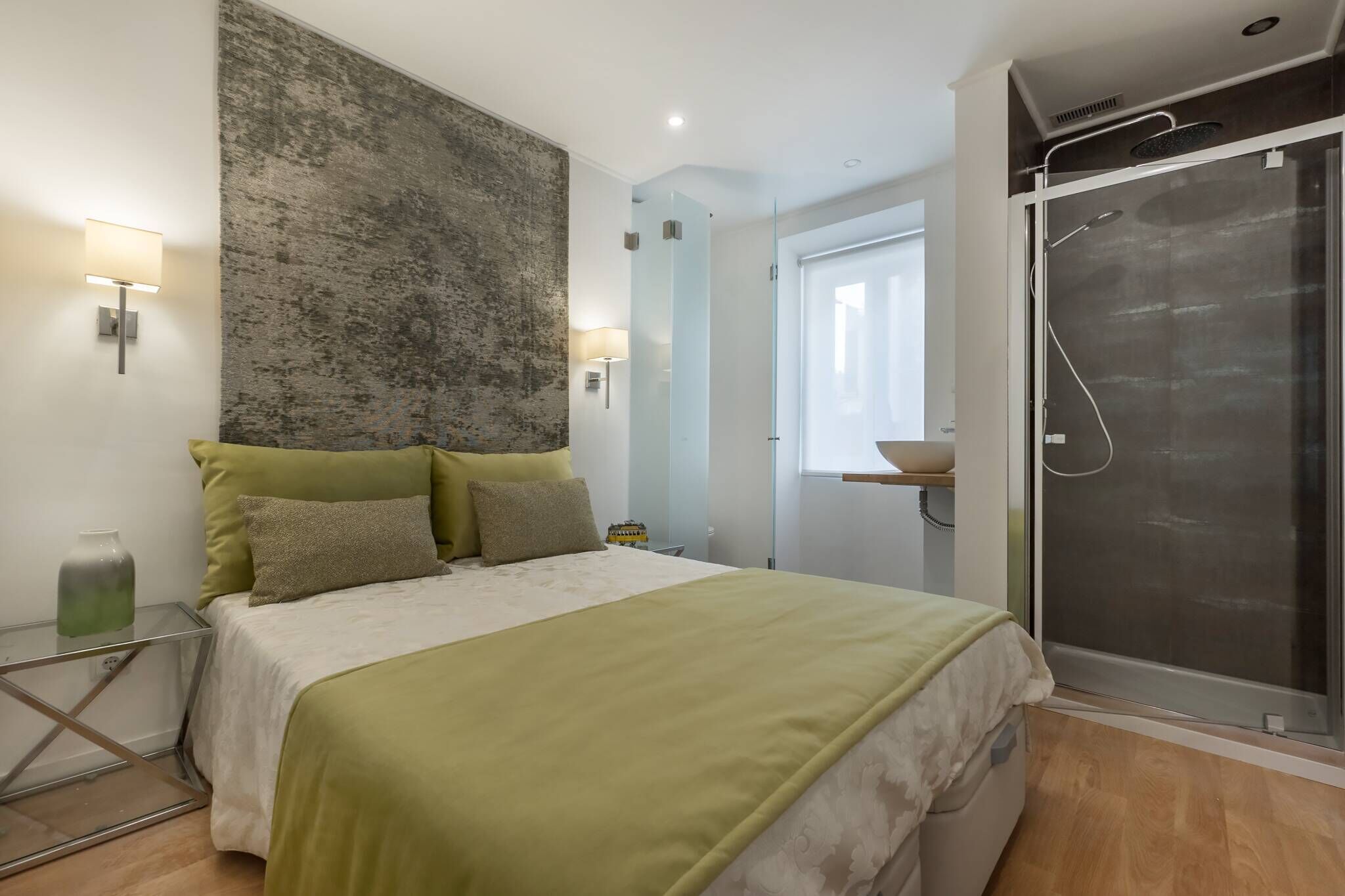 Ein modernes Schlafzimmer mit einem Bett, einer großen strukturierten Wand, einer Duschkabine aus Glas und einem Nachttisch mit Lampe.