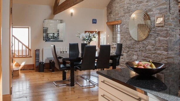 A rustic interior with a stone wall, wooden beams, a dining area with black chairs, a kitchen counter, and a fruit bowl. - Beudy Bach (Llanelli)