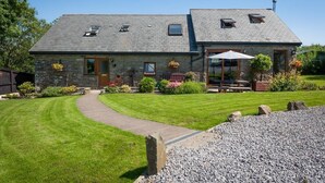 A stone house with a sloped roof, a covered patio, and a well-maintained lawn. - Beudy Bach (Llanelli)