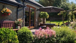 A stone building with a glass door, outdoor seating, and a garden with various plants and flowers. - Beudy Bach (Llanelli)