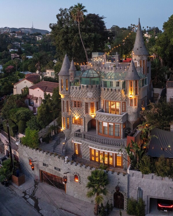 Um edifício grande, com estilo de castelo, com várias torres e ameias, cercado por vegetação exuberante.