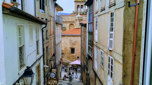 A narrow street with buildings on either side, a bell tower in the distance, and outdoor seating at a café. - ÁgoraLux, The door of Ourense. (Ourense)