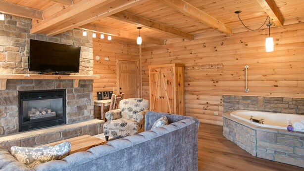 A log cabin interior with a stone fireplace, a television mounted above it, a sofa, a chair, and a bathtub. - Cozy Lodge Fireplace Suite in Ohio's Amish Country (Millersburg)