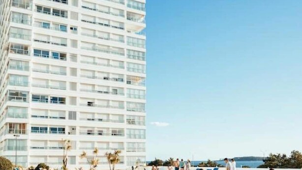 Ein Hotelpoolbereich mit mehreren Balkonebenen, blauem Himmel und Personen, die schwimmen und sich entspannen.