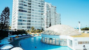 Ein modernes Hotel mit großem Pool, einer kuppelartigen Struktur und Hochhäusern im Hintergrund.