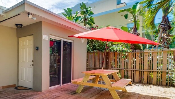 A wooden deck with a picnic table and umbrella, a door, and a small building with a number sign. - 1BR with Private, Fenced Deck (Fort Lauderdale)