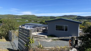 A small, single-story building with a flat roof and a large window, surrounded by a wooden fence and gravel path, overlooking a green landscape with hills. - Modern Bach with a Rural view (Riverton)