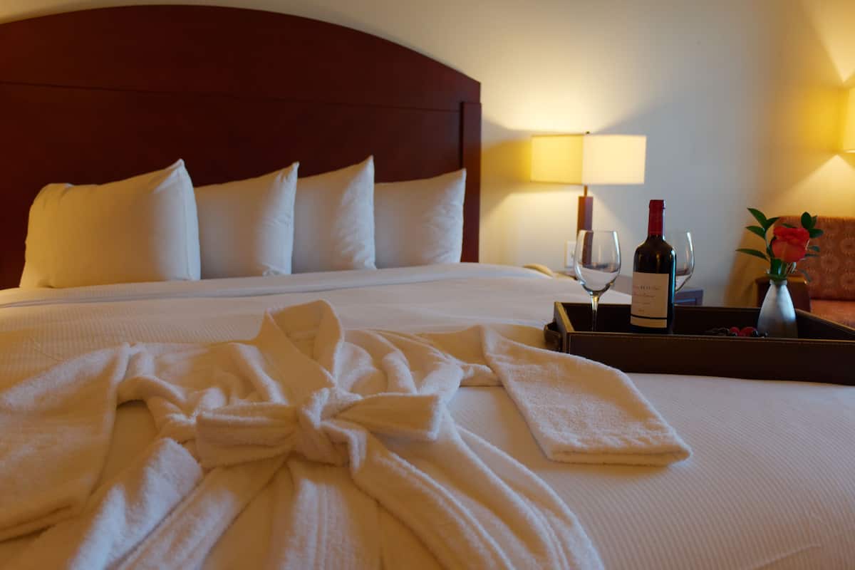Una cama bien hecha con sábanas blancas, una botella de vino, una copa y una bandeja con un jarrón de flores.