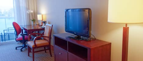 Habitación de hotel con televisión sobre un soporte de madera, un escritorio con una silla, una lámpara y vistas al exterior a través de una ventana.