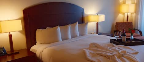 Habitación de hotel con una cama grande, dos lámparas de noche, una silla, una bandeja con una botella y vasos, y un jarrón con una flor.