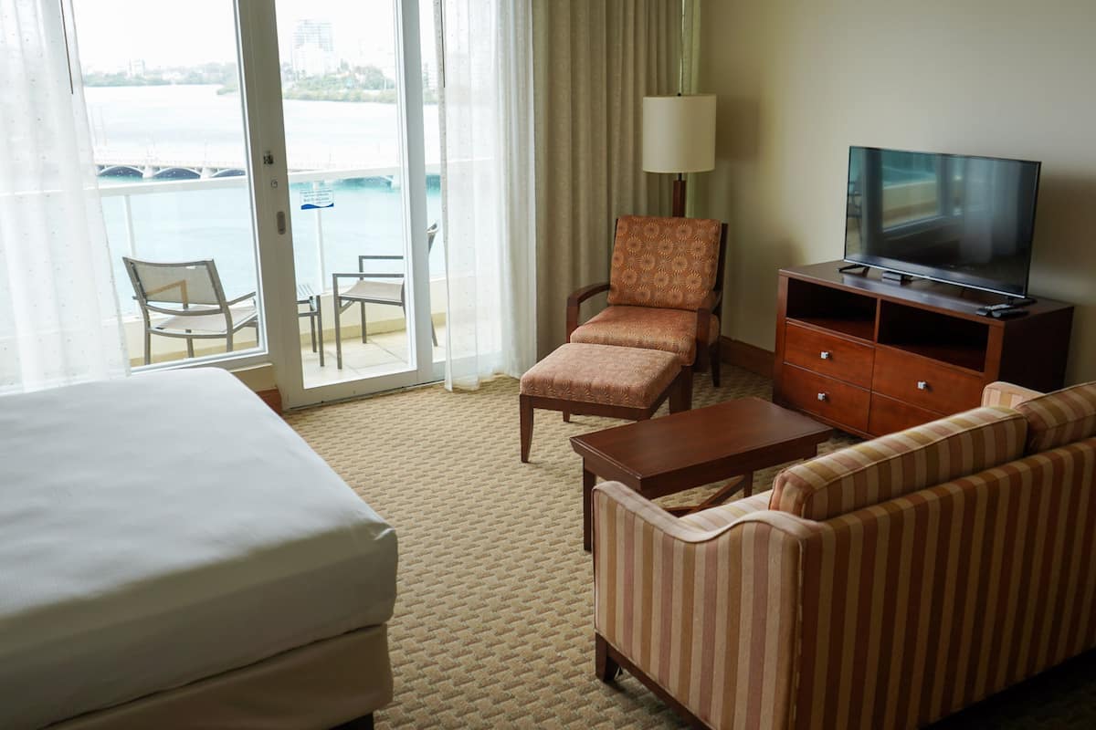 Habitación de hotel con cama, sofá, sillón, televisor sobre un mueble de madera y vista a una masa de agua a través de una puerta de vidrio.