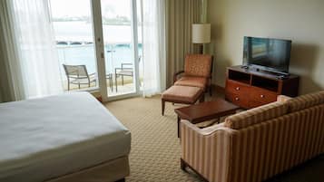 Quarto de hotel com cama, sofá, poltrona, televisão sobre um armário de madeira e vista para a água através de uma porta de vidro.