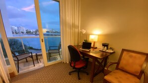 Quarto de hotel com escrivaninha, laptop, cadeira, luminária e vista para a paisagem urbana e para a água.