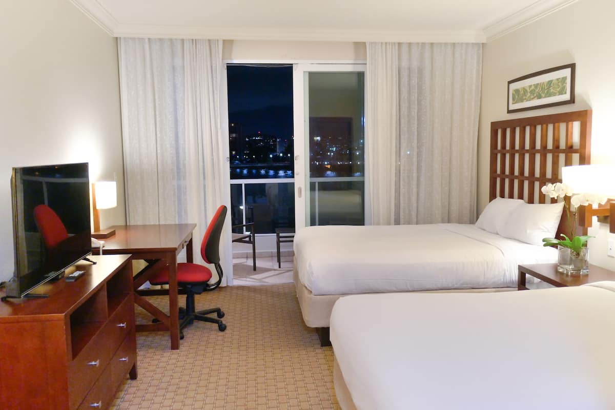 Habitación de hotel con televisión, escritorio, dos camas, una silla y vista a un paisaje urbano de noche.