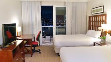 A hotel room with a TV, desk, two beds, a chair, and a view of a cityscape at night.