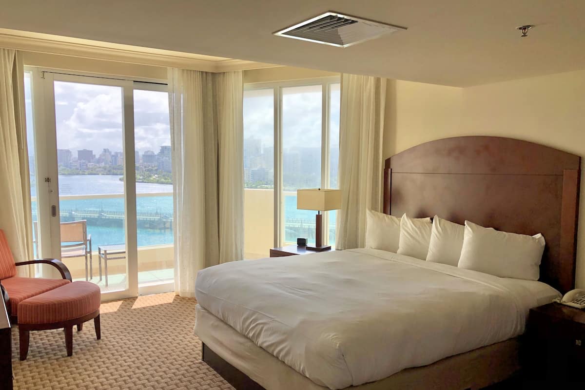 Habitación de hotel con una cama grande, una silla, una mesita de noche y vistas a la ciudad y al agua.