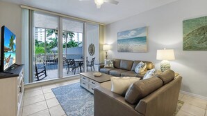 Sala de estar con sofá, mesa de centro, televisor y un balcón con vistas a palmeras y edificios.