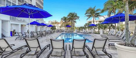A poolside area with lounge chairs and blue umbrellas, a clear sky, and a building in the background.