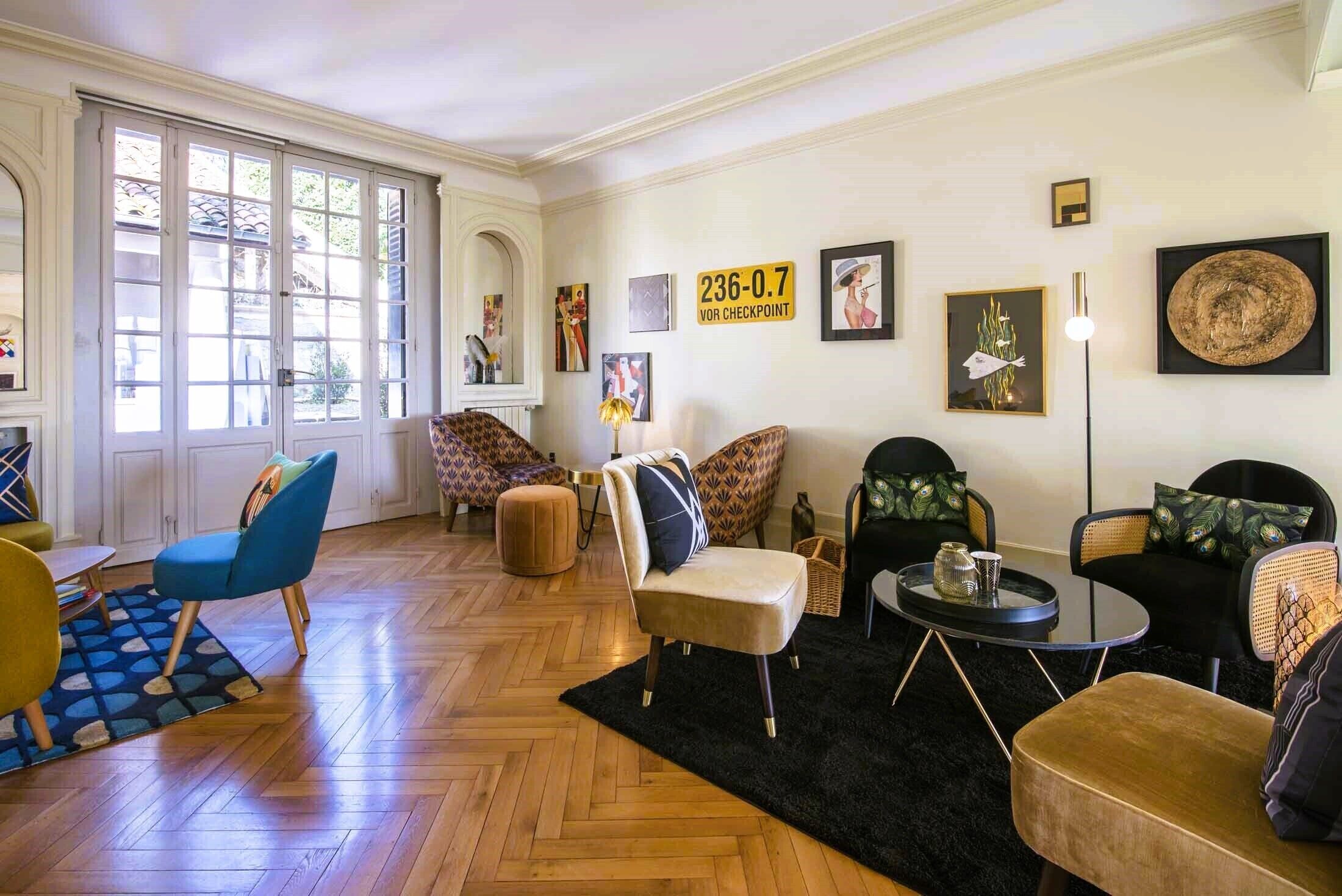 Ein geräumiges Wohnzimmer mit großem Fenster, einem schwarzen Teppich und verschiedenen Sitzgelegenheiten, darunter ein blauer Sessel und ein beiges Sofa.