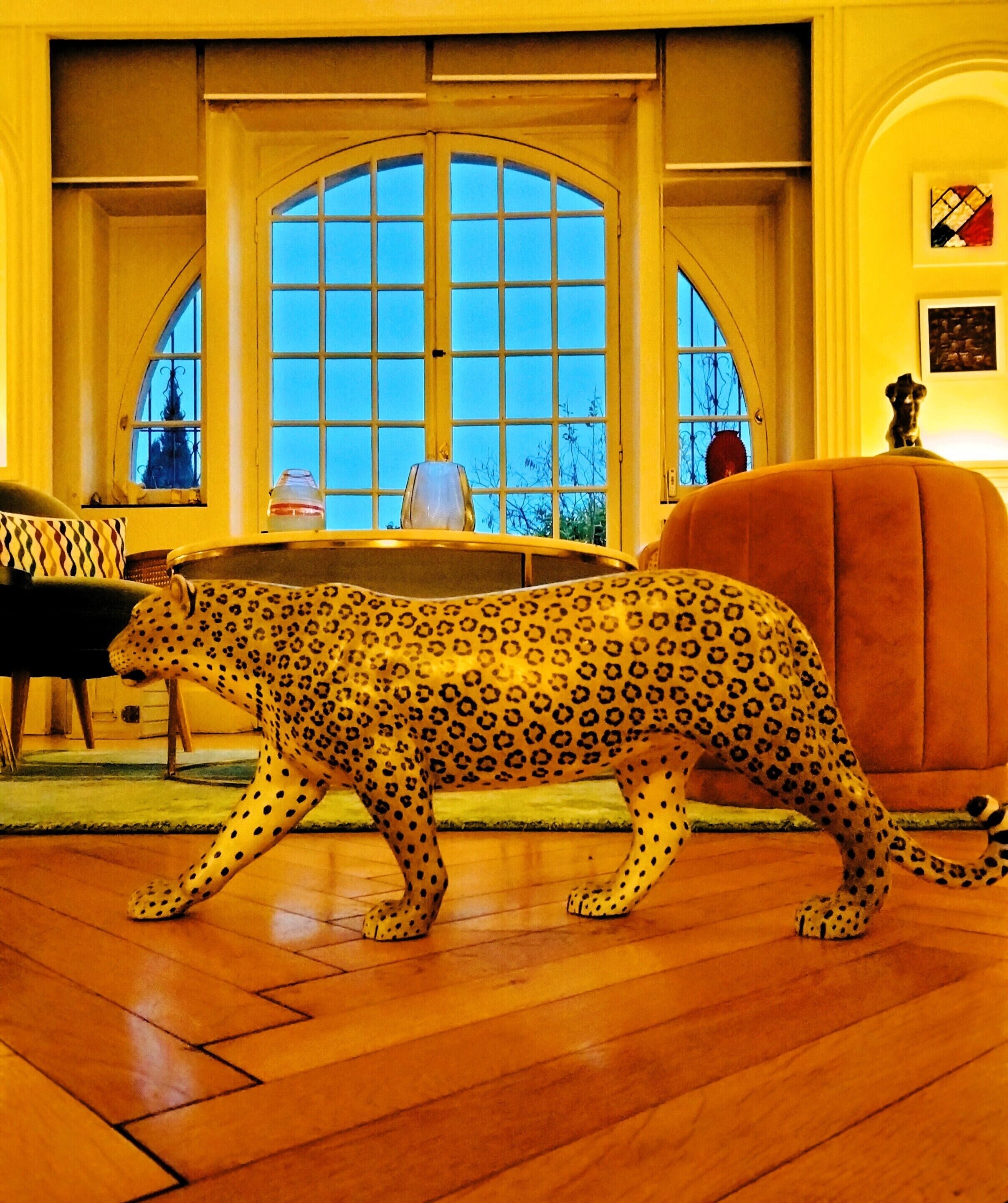 Una estatua de leopardo en una habitación con suelo de madera, un ventanal arqueado grande y una silla.