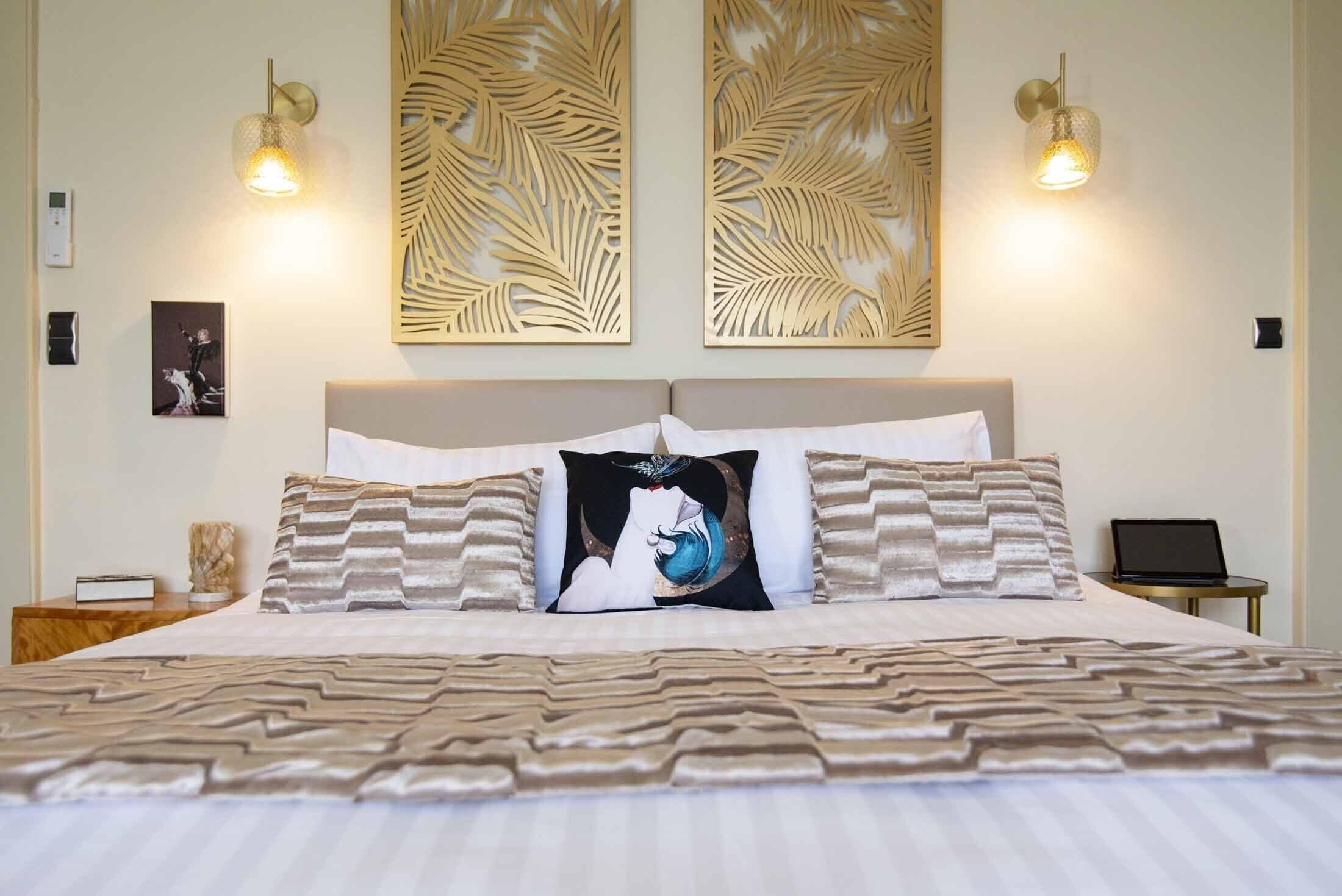 Una cama bien tendida con ropa de cama estampada, dos lámparas de pared y un ordenador portátil sobre una mesita de noche.