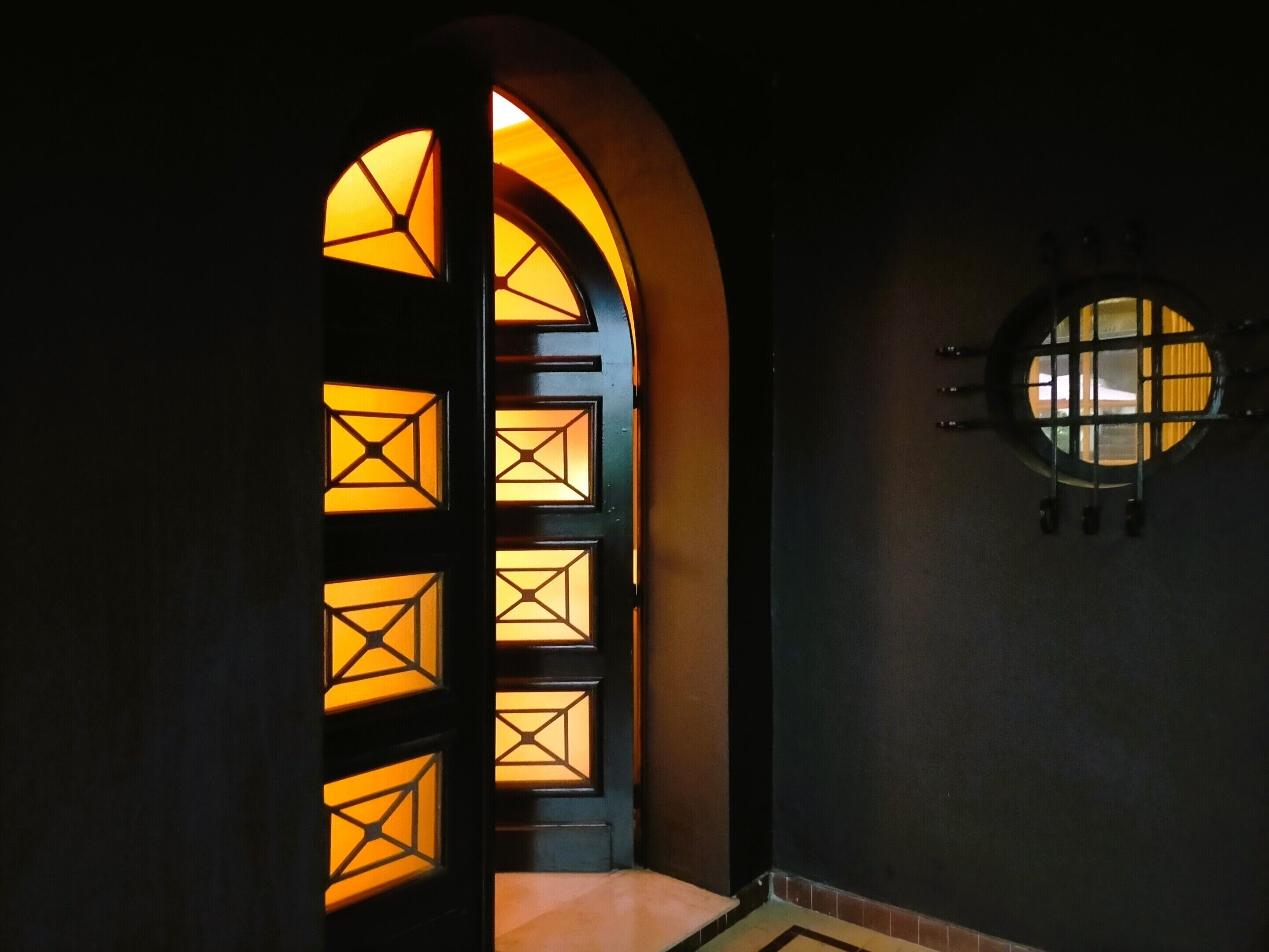 Un pasillo oscuro con una puerta que presenta diseños geométricos iluminados y un ventanal redondo.