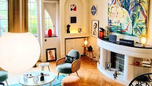 A spacious living room with a fireplace, a large painting, and a modern chandelier. - Quand la Ville Dort (Agen)