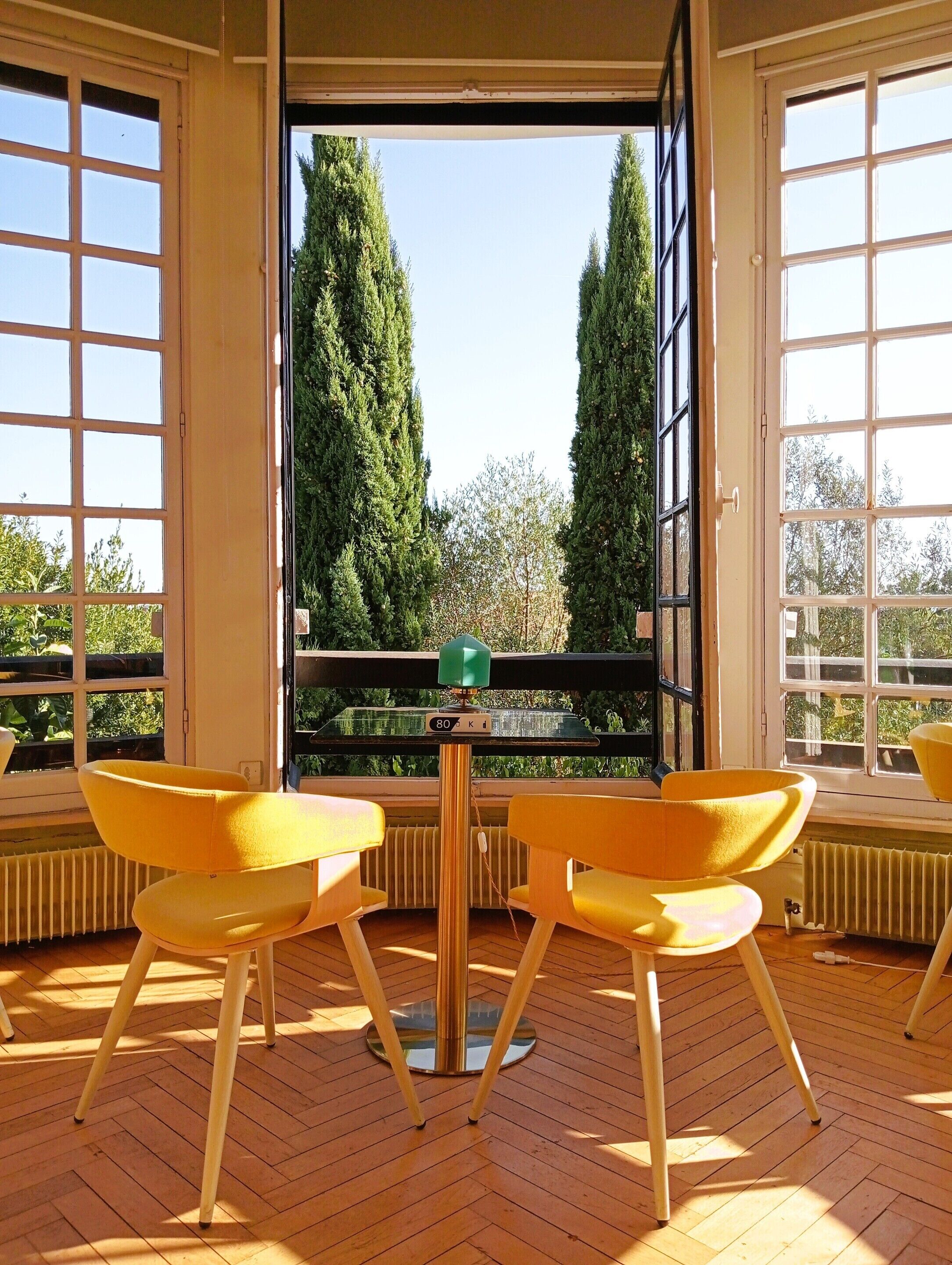 A room with a wooden floor, two yellow chairs, a small table, and large windows with a view of tall trees.