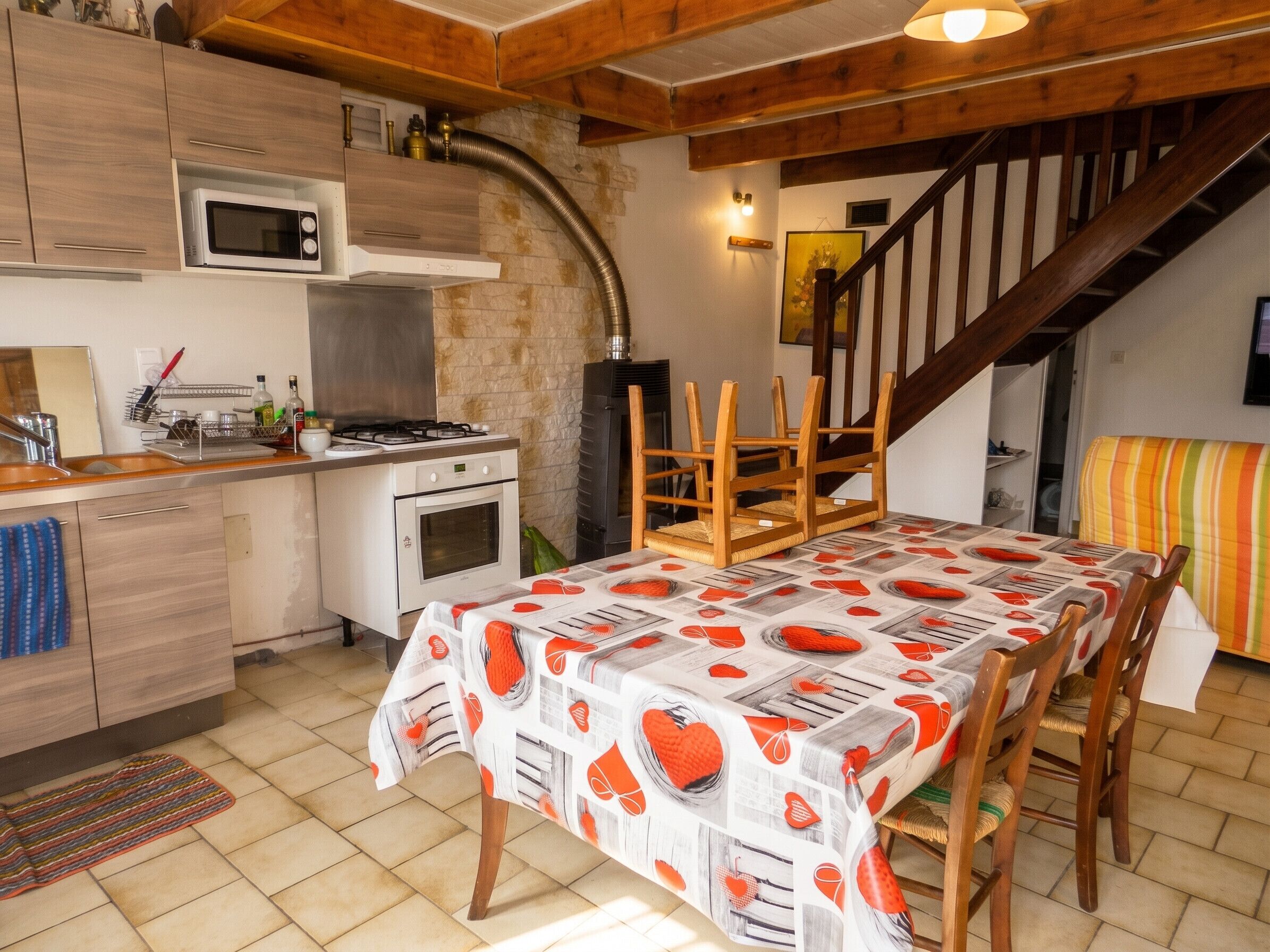 A kitchen with a microwave, oven, and gas stove, wooden chairs, a table with a patterned tablecloth, and a staircase leading to a loft.