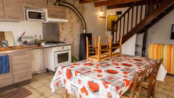 A kitchen with a microwave, oven, and gas stove, wooden chairs, a table with a patterned tablecloth, and a staircase leading to a loft.
