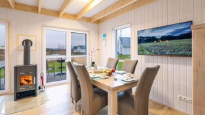A cozy room with a wooden ceiling, a fireplace, a dining table with chairs, and a large window with a view of a field and a house. - Gorgeous home in OstseeResort Olpenitz with sauna (OstseeResort Olpenitz)
