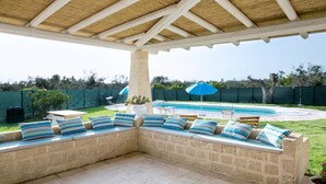 En overdækket terrasse med stenbænke og en pool i baggrunden.