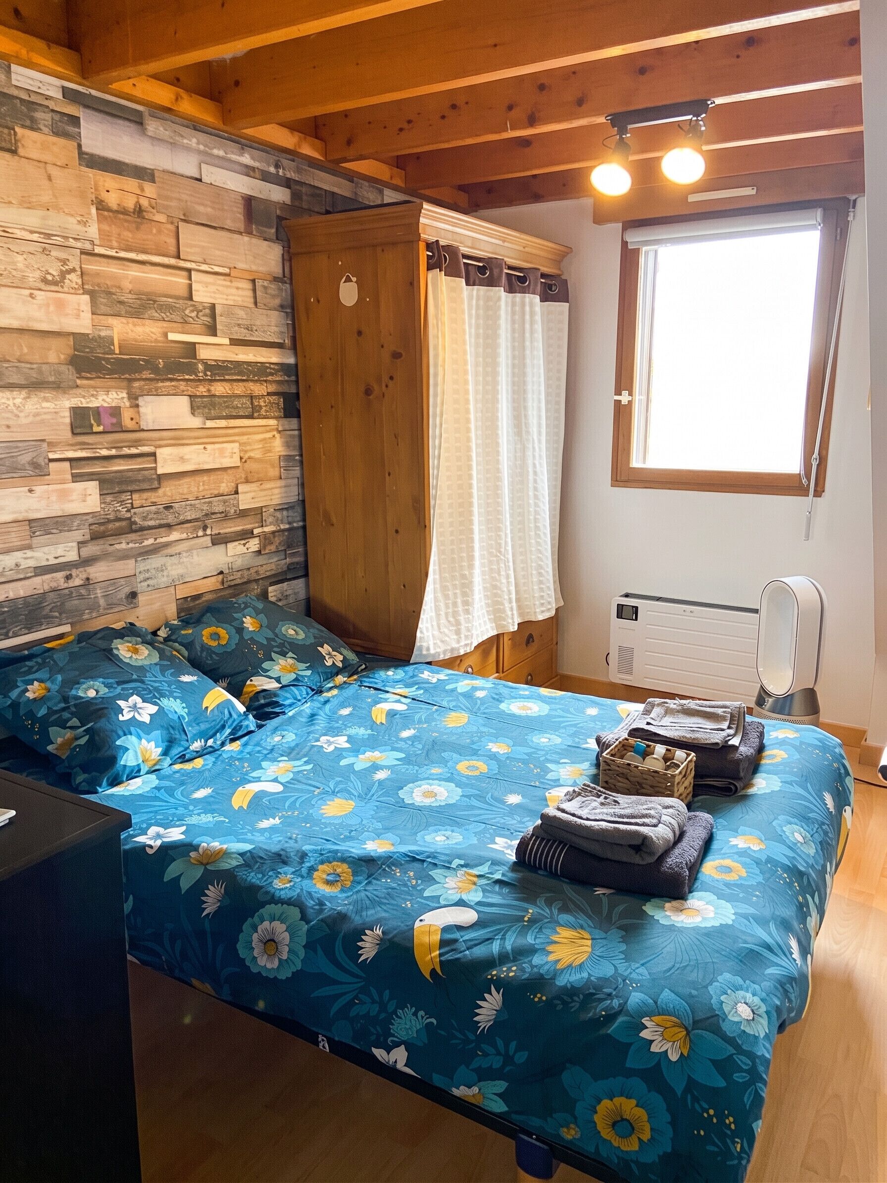 A bedroom with a wooden ceiling, a bed with a floral bedspread, a wooden wardrobe, a window with curtains, and a radiator.