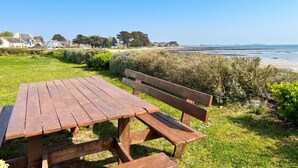 Une table et un banc de pique-nique en bois, installés dans un espace herbeux, offrent une vue sur la plage et les maisons qui se trouvent au loin.
