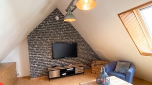 Une pièce de style « loft » avec un plafond en pente, un mur en briques apparentes, un téléviseur à écran plat fixé au mur, une table basse en bois, un fauteuil bleu et un pouf jaune.