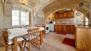 A rustic stone kitchen with wooden cabinets, a dining table set for four, and a red rug. - Discover Tranquility: rustic elegance in this beautifully converted farmhouse. (San Lawrenz)