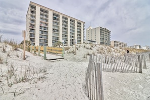 A beachfront hotel with multiple floors, a sandy beach in the foreground, and a wooden fence.