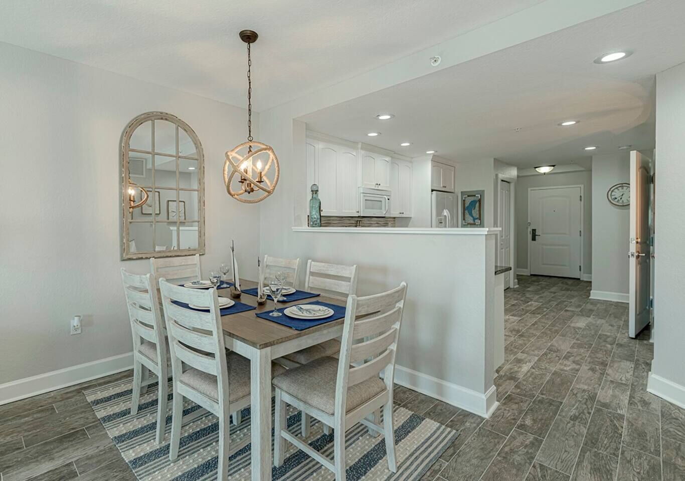 A dining area with a wooden table and white chairs, a kitchen with white cabinets, and a hallway leading to a door.